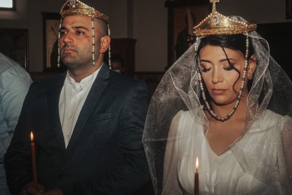 Bride and groom in traditional wedding attire with crowns, holding candles in solemn ceremony.